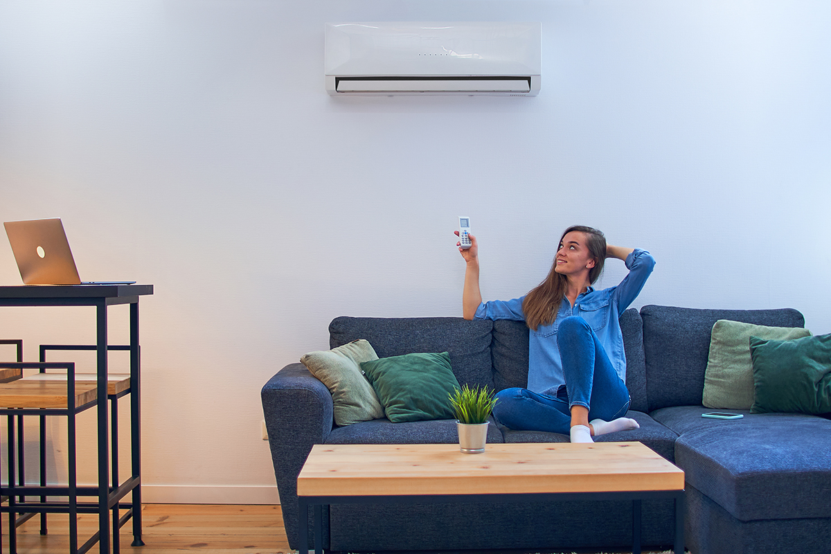 Young happy woman sitting on couch under air conditioner and adjusting comfort temperature with remote control at modern home