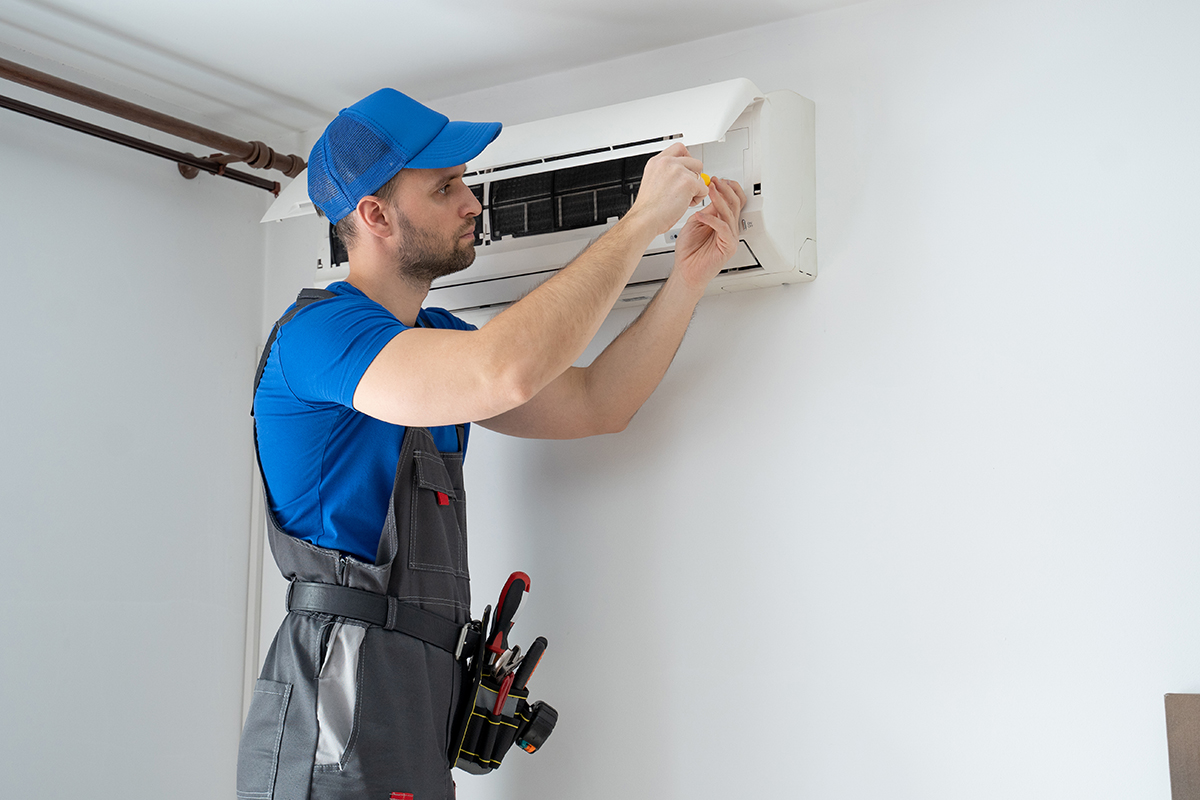 Male technician in overalls and a blue cap repairs an air conditioner on the wall.